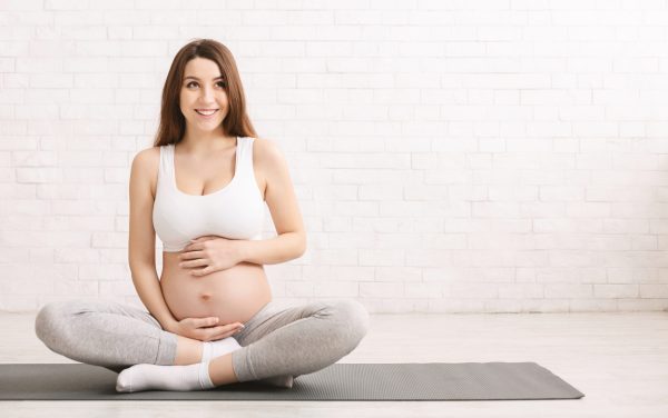 Active pregnant woman touching her belly, resting after workout on fitness mat, looking at empty space